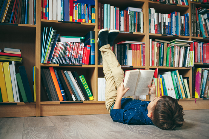 A boy lying on the ground on his back reading a book with his feet up against the bookshelves